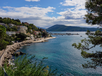 This landscape photograph captures a scenic view looking towards Herceg Novi from the Mirko Komnenović museum gardens, during a bright spring morning in Montenegro. The image prominently features the charming coastal town of Herceg Novi extending along the Adriatic shoreline, with a sweep of colourful buildings and lush greenery along the popular Pet Danica promenade. In the background, the hills of Boka Bay rise gently, while the clear blue waters of the bay are dotted with moored boats and a distant marina, highlighting the picturesque appeal of this part of Montenegro. The photo's vibrant lighting and tranquil atmosphere evoke the serene beauty for which Boka Bay and Herceg Novi are renowned.